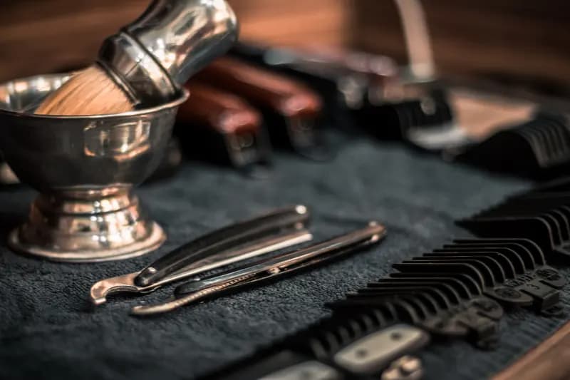 Pincel de barba, tigela de metal, navalhas e pentes de máquina sobre uma toalha escura